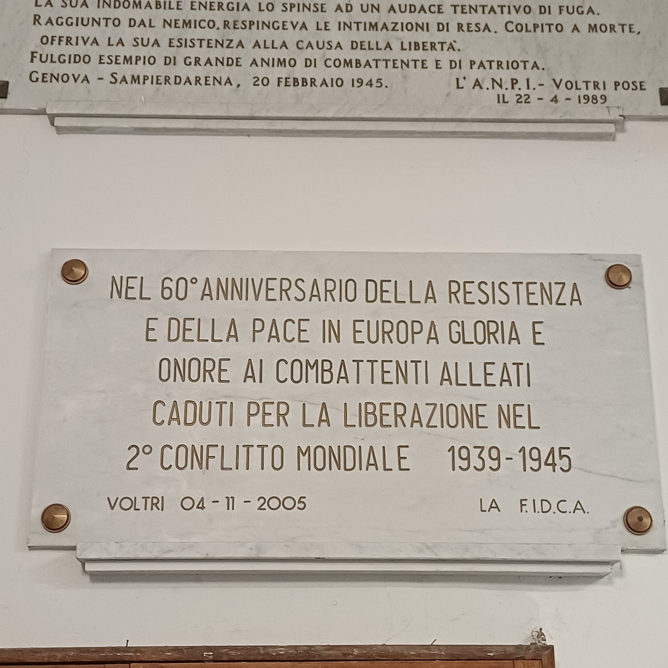 Lapide in marmo con iscrizione, fissata a parete con quattro borchie metalliche angolari.