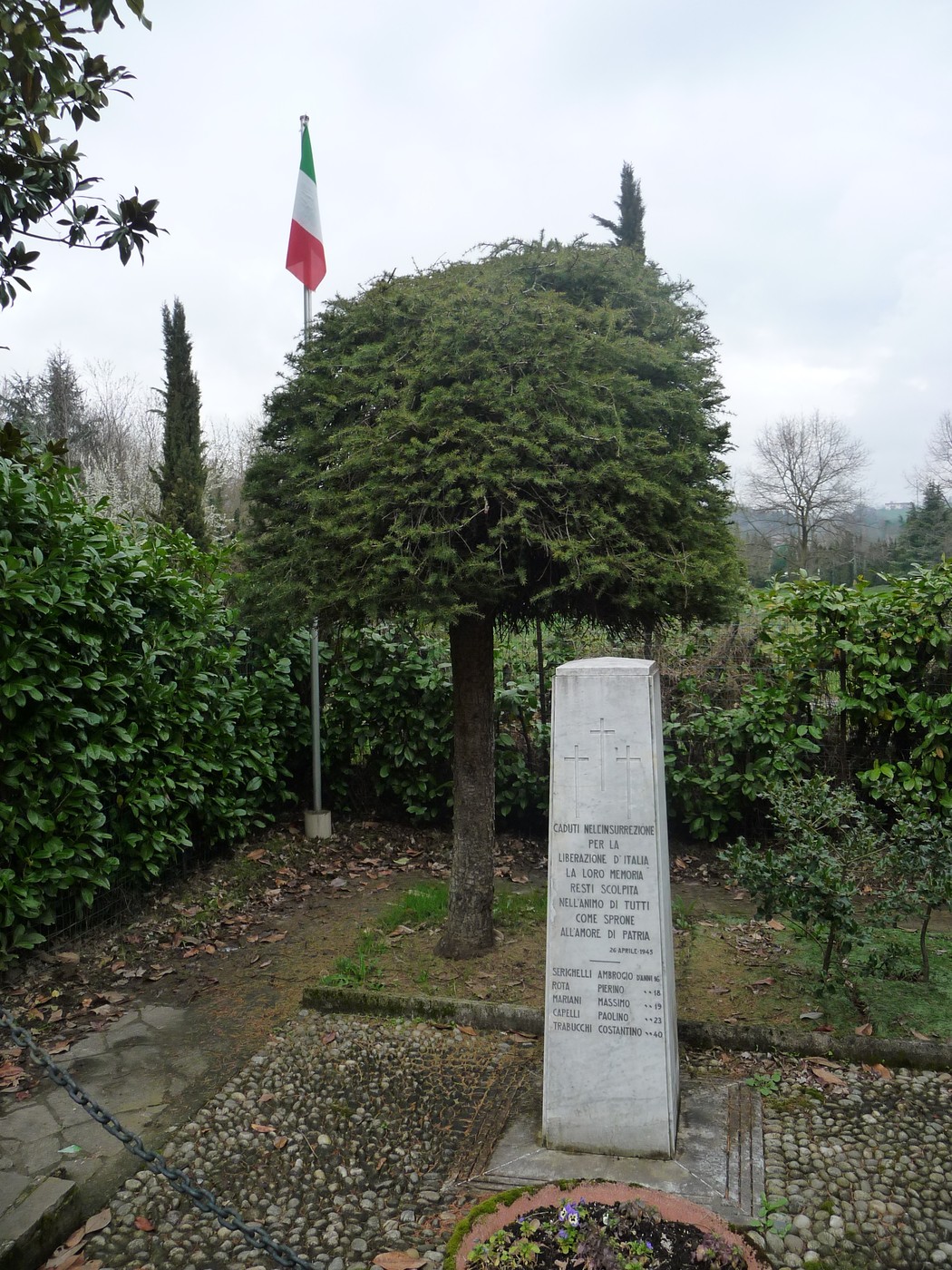 Cippo commemorativo ai caduti partigiani di Longuelo, circondato da vegetazione e con bandiera italiana.
