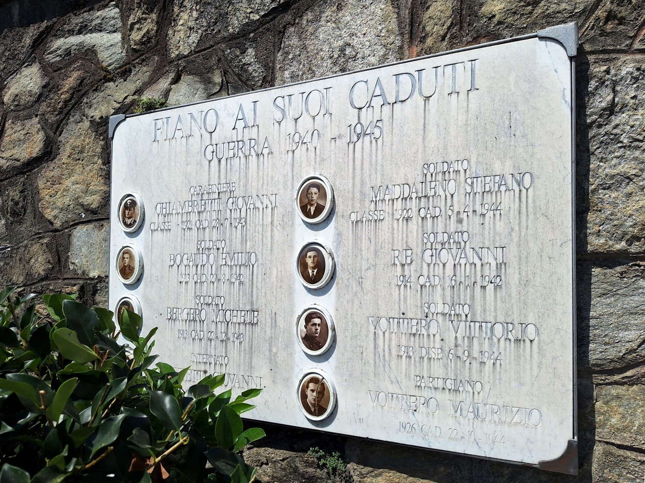 Lapide commemorativa dedicata ai caduti della guerra 1940-1945 con nomi e fotografie. Ambientazione rustica.