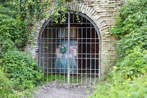 Ingresso con cancello metallico in una struttura storica, circondato da vegetazione e muri in mattoni.