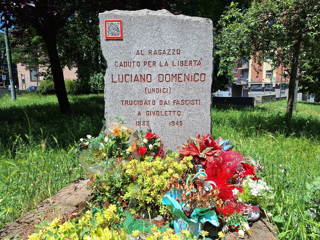 Cippo commemorativo dedicato a Luciano Domenico, con fiori e tributi per il suo sacrificio durante la guerra.