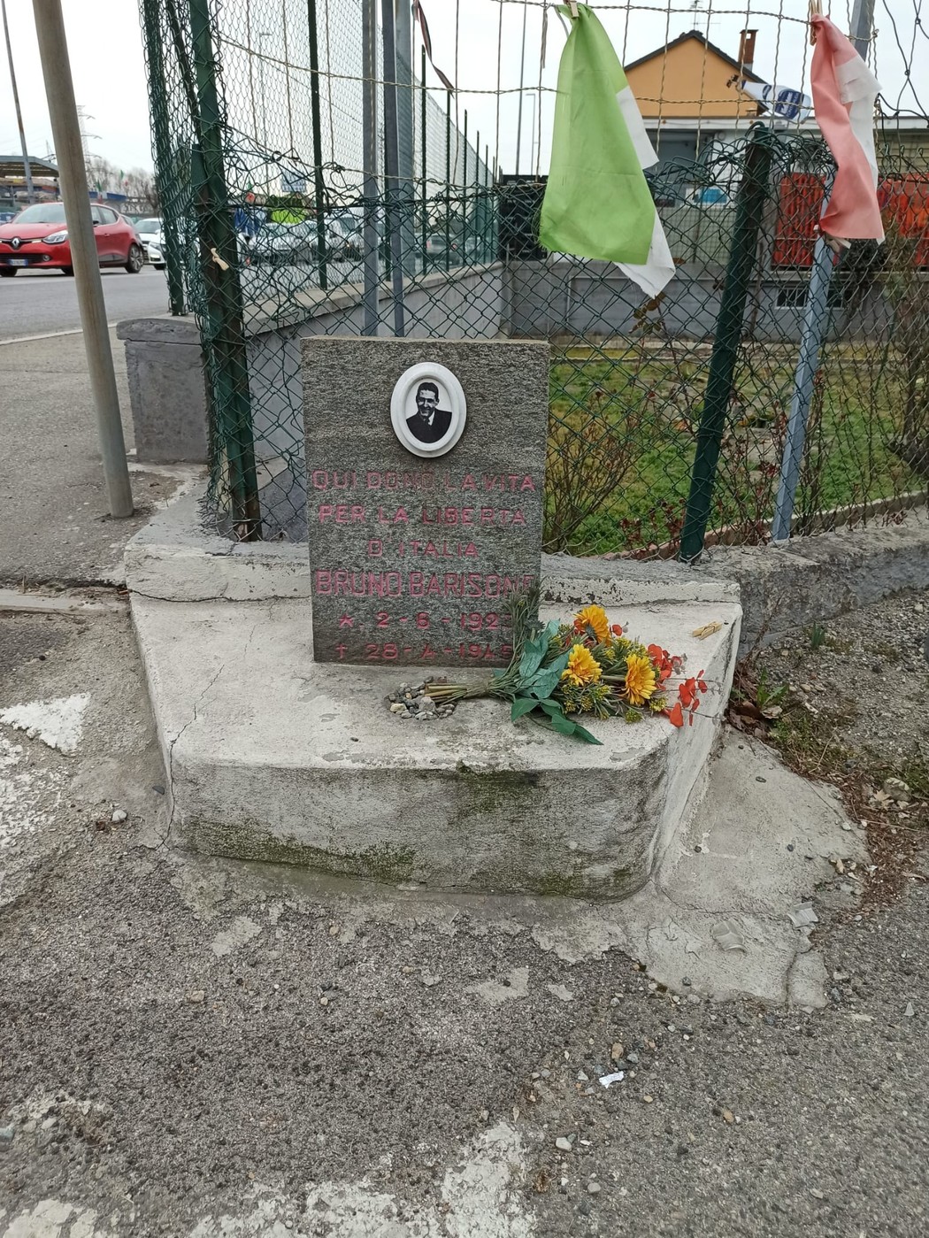 Cippo commemorativo dedicato a Barisone, con fiori e bandiere, situato all'incrocio di una strada.