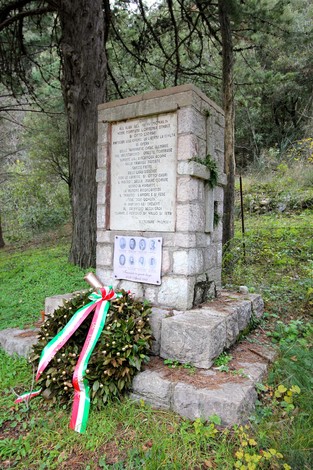 Monumento commemorativo con lapide in pietra e corona di alloro, immerso nel verde di un bosco.