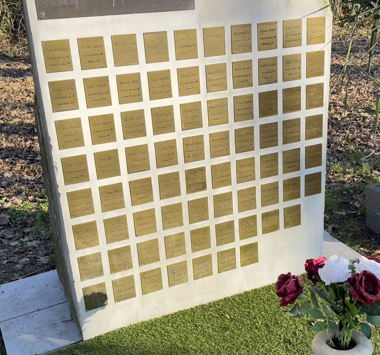 Il Giardino della Memoria con lastre commemorative su una parete, circondato da vegetazione e fiori.