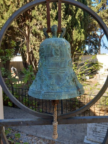 Campana bronzea al "Sacrario dei caduti di Kos", circondata da un giardino, simbolo di memoria e onore.