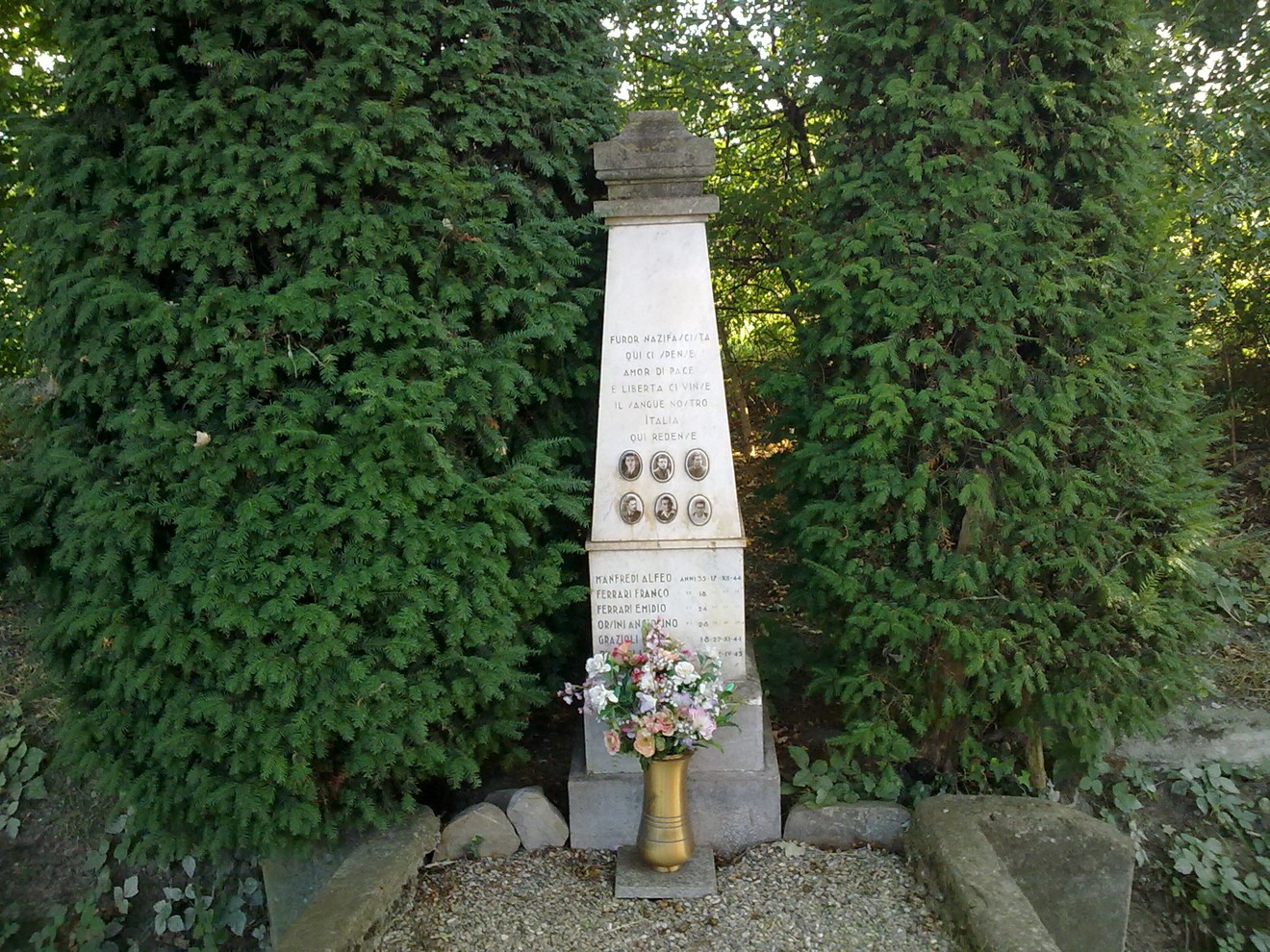 Monumento commemorativo a Manfredi e altri, circondato da alberi e decorato con fiori.