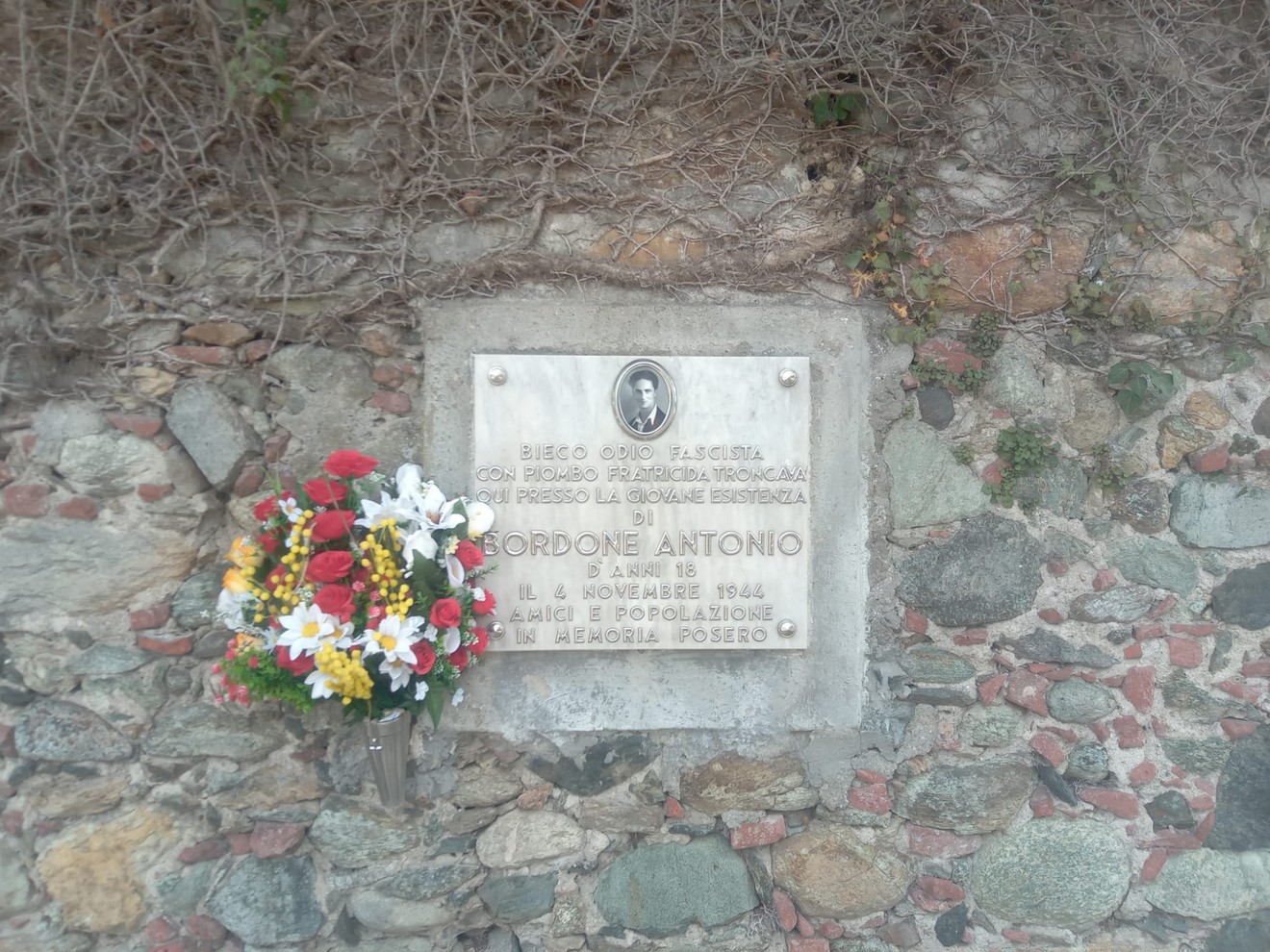 Lapide commemorativa su un muro di pietra, circondata da fiori colorati, in memoria di Bordone Antonio.