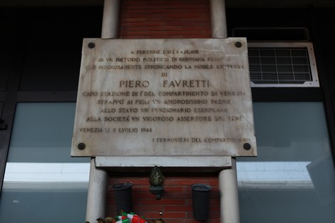 Lapide commemorativa a Piero Favretti, situata in un contesto urbano, con dettagli architettonici circostanti.