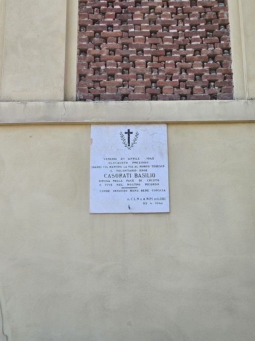 Lapide in marmo con iscrizione, sormontata da una croce, affissa su parete esterna sotto una finestra in mattoni.