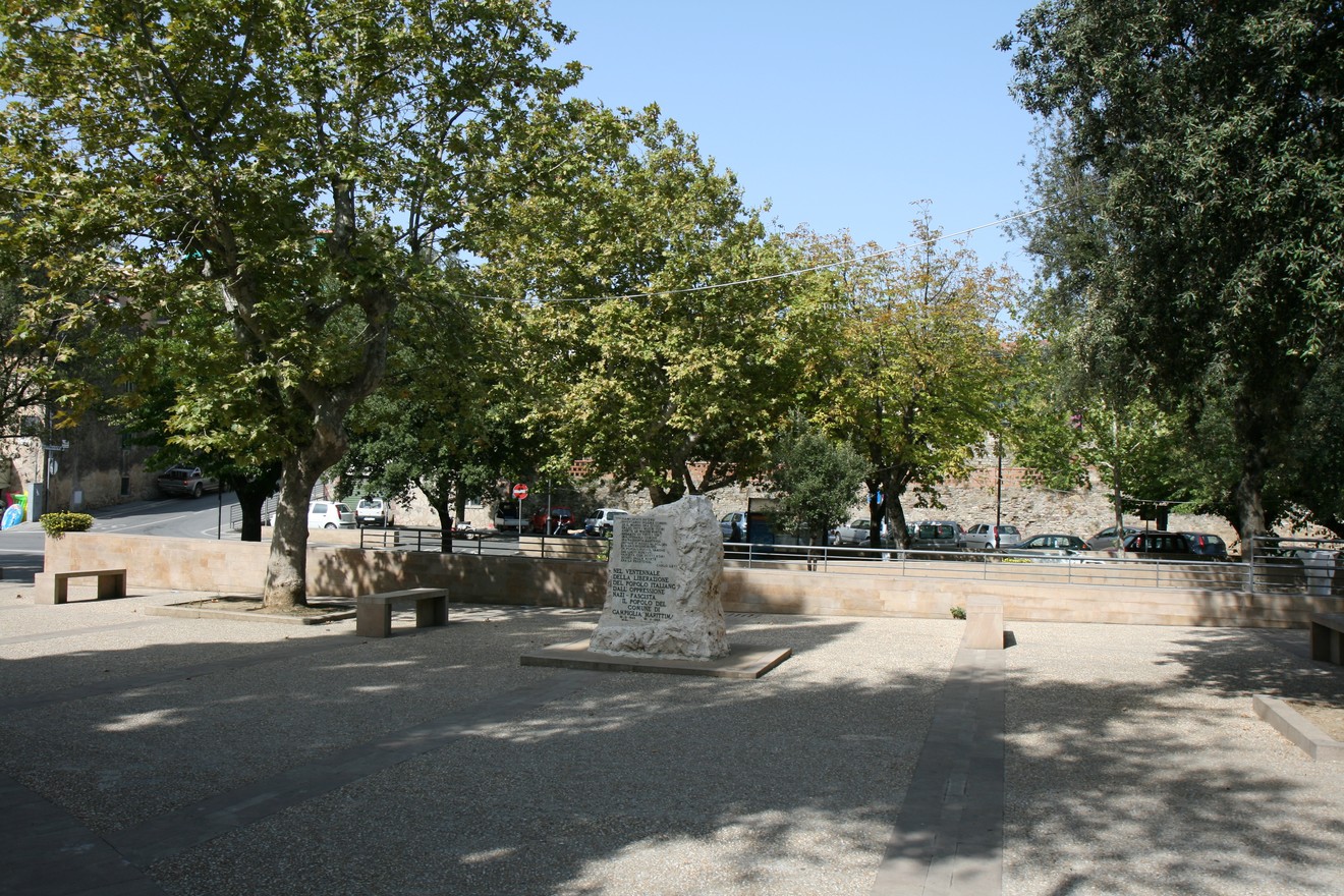 Monumento del ventennale immerso in un'area verde con alberi e panchine circostanti.