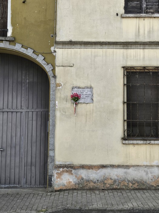 Lapide commemorativa affissa su una parete esterna, con fiori decorativi nei pressi.