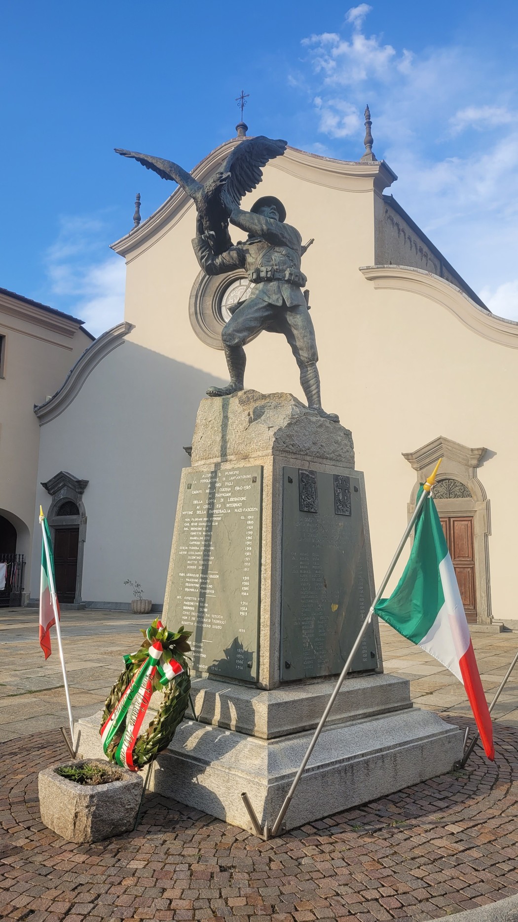 Monumento in bronzo con soldato e aquila su basamento di pietra con iscrizione, bandiere italiane e corona ai lati.