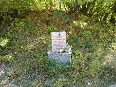 Cippo commemorativo dedicato a Rinaldi, circondato da natura e omaggi floreali.