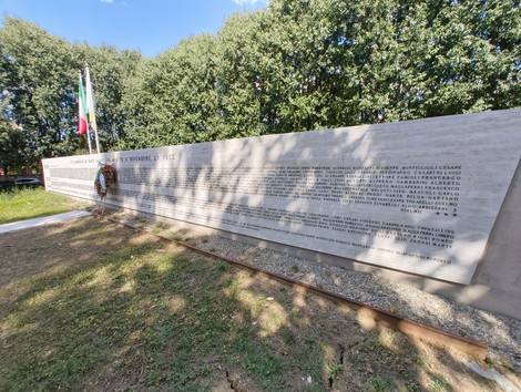 Monumento ai Caduti con iscrizioni commemorative, decorato da una corona e bandiere. Spazio verde circostante.
