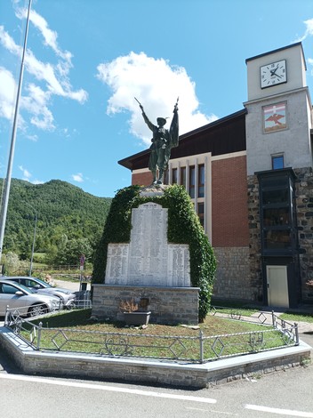 Monumento ai caduti con statua alato, circondato da vegetazione e edifici, in un paesaggio montano.