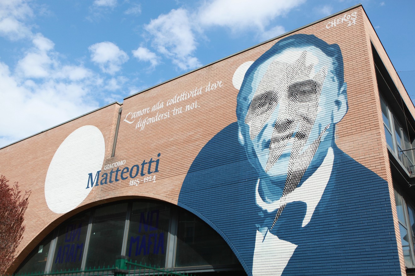 Murale dedicato a Giacomo Matteotti, raffigurante il suo volto sopra un edificio, con sfondo blu e nuvole.