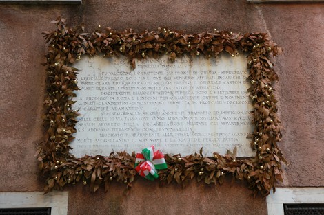 Lapide commemorativa adornata con foglie e un nastro tricolore su una parete.