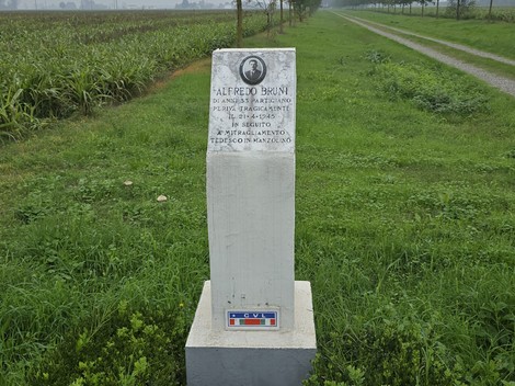 Cippo commemorativo dedicato a Alfredo Bruni, situato lungo una strada circondata da campi verdi.