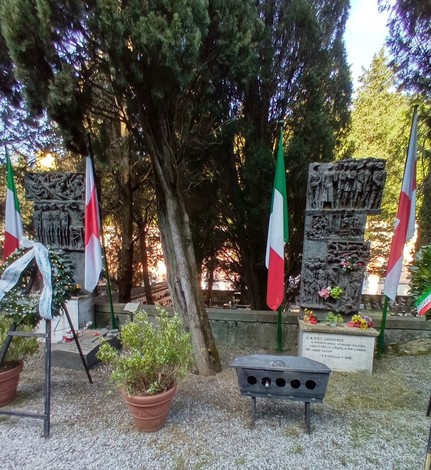 Monumento ai deportati con sculture e bandiere italiane, immerso in una cornice di alberi e piante.