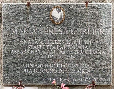 Lapide in marmo grigio con ritratto fotografico ovale e iscrizione, fiori deposti alla base.
