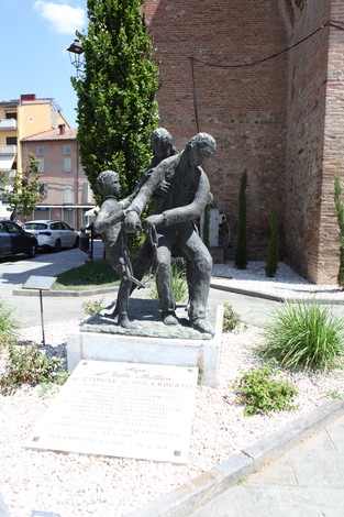 Monumento ai Caduti con una scultura di uomini e bambini in una piazza, circondato da vegetazione e strade.