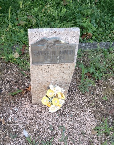 Cippo commemorativo a Ravanelli, decorato con fiori, immerso in un'area di verde.