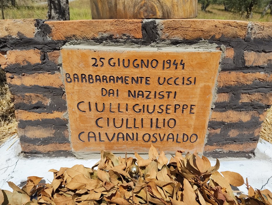 Cippo in mattoni con lapide di marmo incisa, decorato con corone di foglie secche alla base.