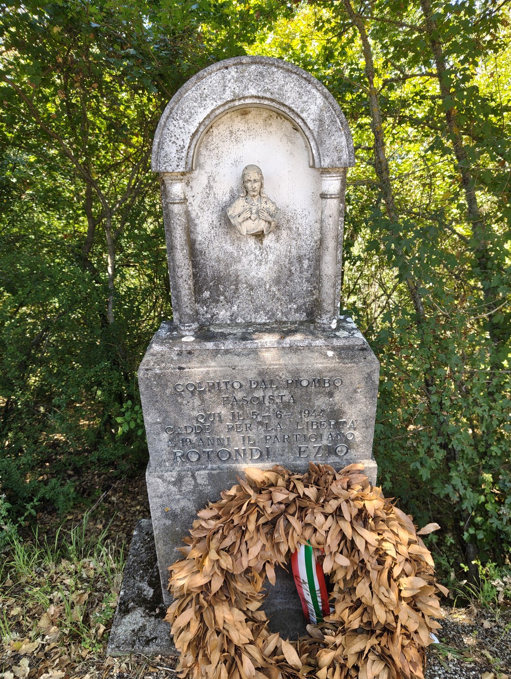 Cippo in pietra con bassorilievo di una figura e iscrizione, corona di alloro ai piedi, nel bosco.