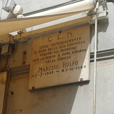 Lapide marmorea con iscrizione dedicata a Narciso Rulfo, fissata su parete esterna sotto una tenda.