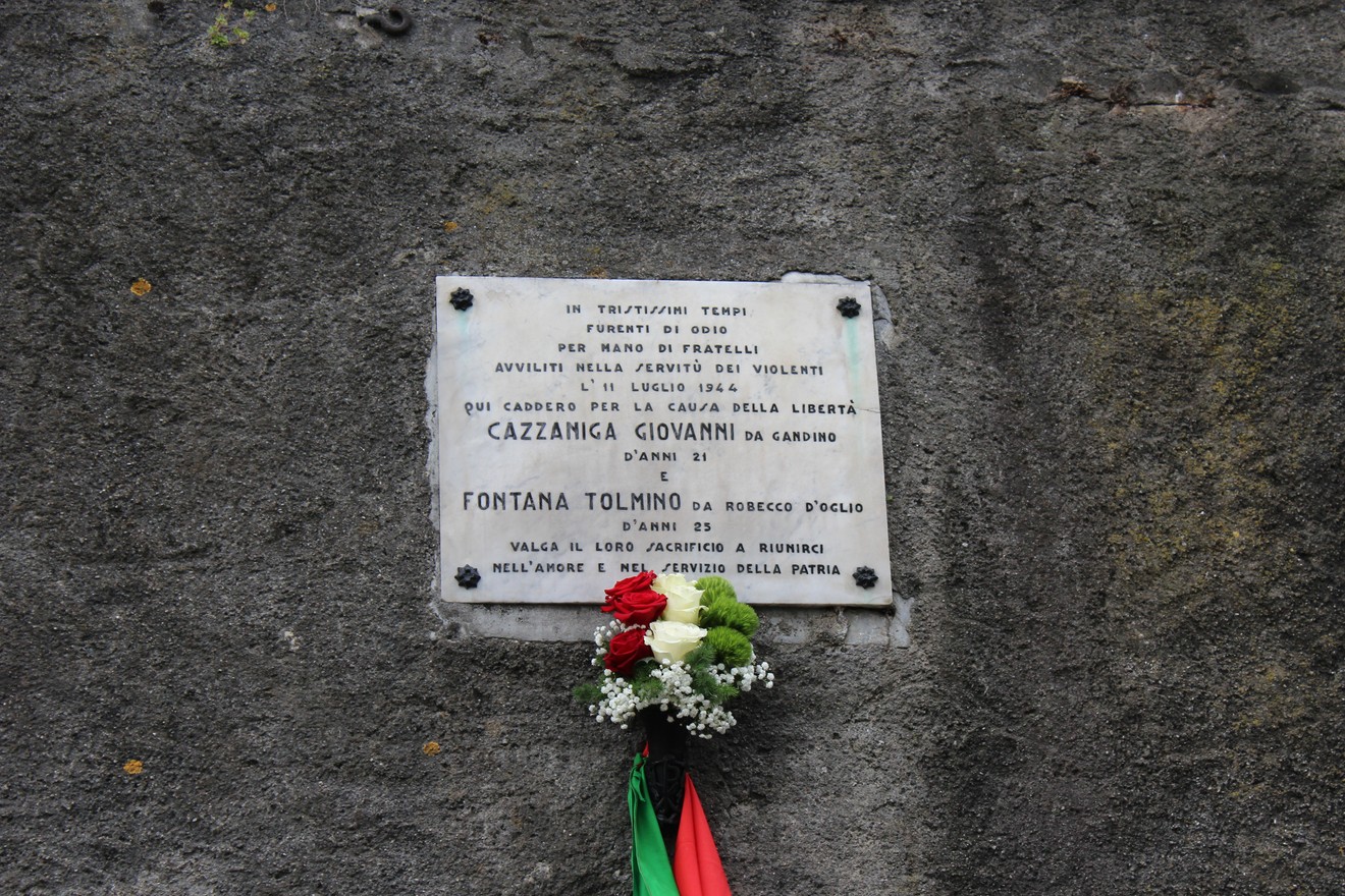 Lapide commemorativa dedicata a Cazzaniga e Fontana, con decorazioni floreali, su una parete di pietra.