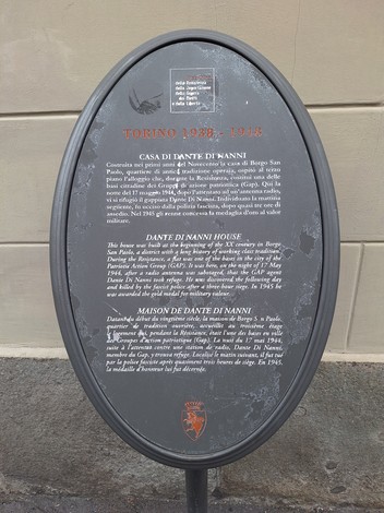 Targa ovale in metallo scuro con iscrizione multilingue, montata su palo su una strada.