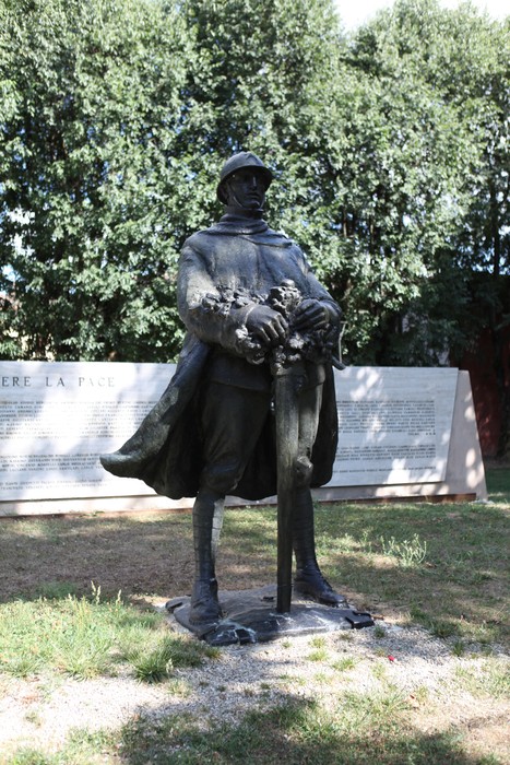 Monumento ai Caduti con statua di soldato in uniforme, circondato da vegetazione e pannelli informativi.