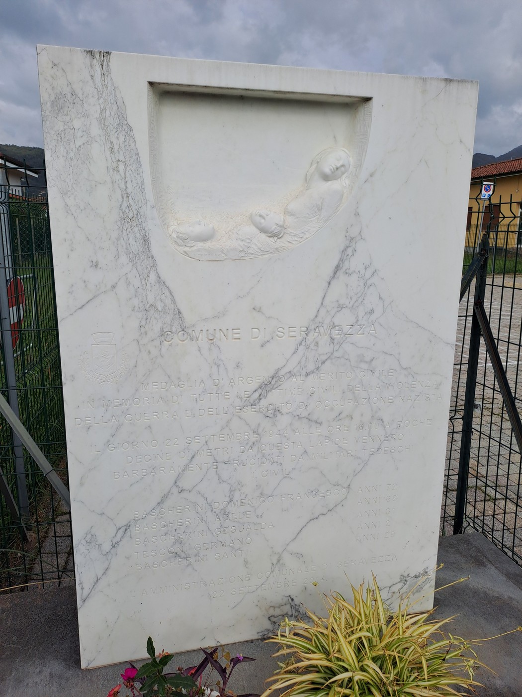 Lapide in marmo bianco con bassorilievo di colombe e iscrizione, all'aperto dietro una recinzione.