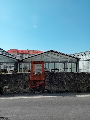 Lapide in pietra visibile accanto a serra, con edifici residenziali sullo sfondo e cielo sereno.