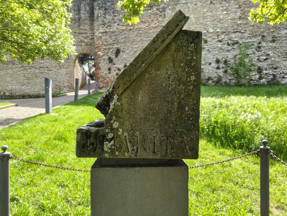 Cippo a Meuret immerso nel verde, con rovine storiche sullo sfondo e dettagli di usura sulla pietra.