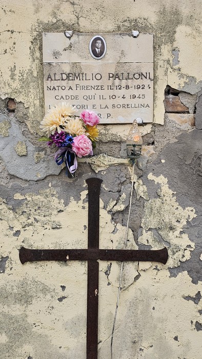 Croce in ferro con ai piedi fiori e una foto affissa su un muro degradato.