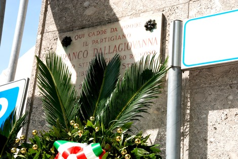 Lapide commemorativa di Franco Dellagiovanna, partigiano, con decorazioni floreali e segnaletica stradale.