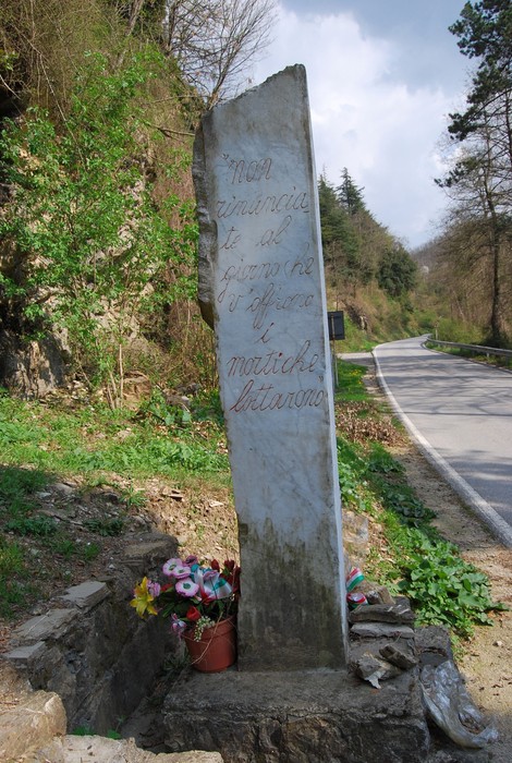 Monumento a Damico, un pilastro in pietra con decorazioni floreali e un paesaggio naturale circostante.