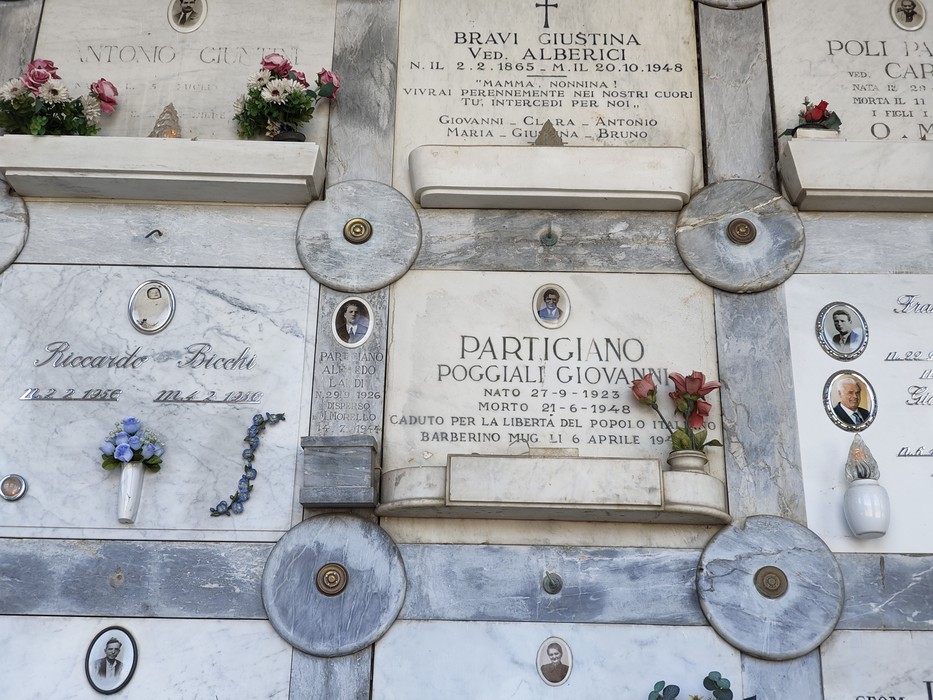 Parete di loculi in marmo con lapidi e foto ovali, al centro epigrafe dedicata a un partigiano.