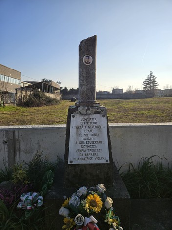 Cippo commemorativo a Guatelli, con fiori e dedica in memoria di una giovane scomparsa. Sullo sfondo, edifici e un cielo sereno.