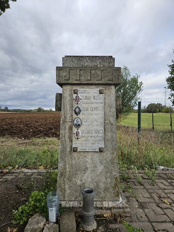Cippo commemorativo dedicato a Corradi, Olmi, Verti e Dall’Aglio, circondato da un paesaggio rurale.