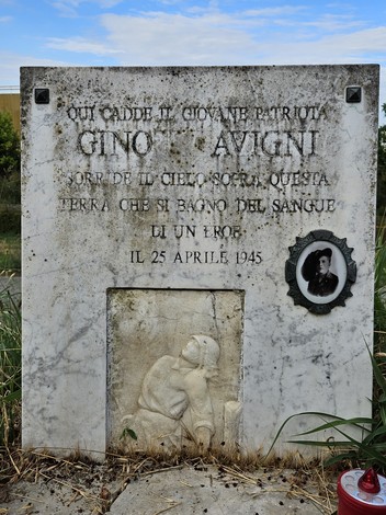 Cippo commemorativo in onore di Gino Avigni, giovane patriota caduto il 25 aprile 1945, circondato da vegetazione.