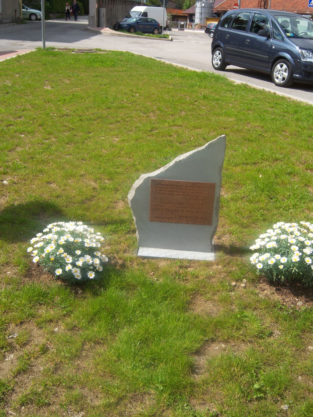 Cippo commemorativo circondato da fiori, situato in un'area verde con automobili in lontananza.