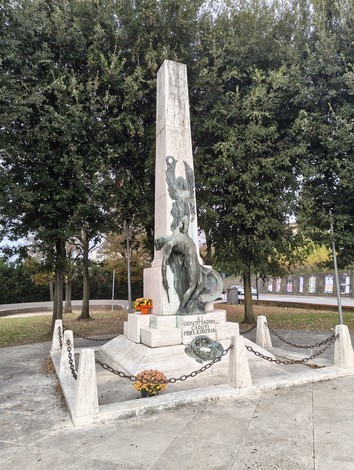 Monumento ai caduti con statue commemorative e decorazioni floreali, circondato da alberi e catene.