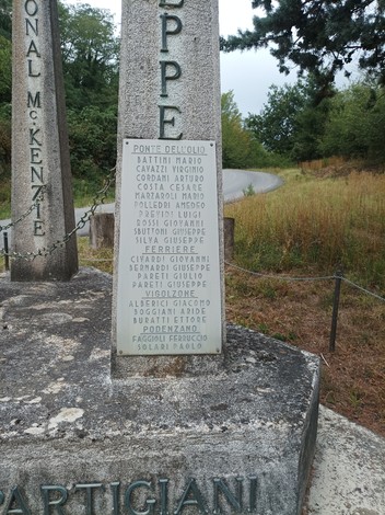Monumento ai partigiani della Valnure, dedicato ai caduti con nomi incisi su pietra. Verde circostante e strada in lontananza.