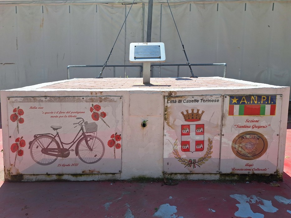 Targa decorativa a Noce con simboli locali e un'immagine di bicicletta in un contesto urbano.