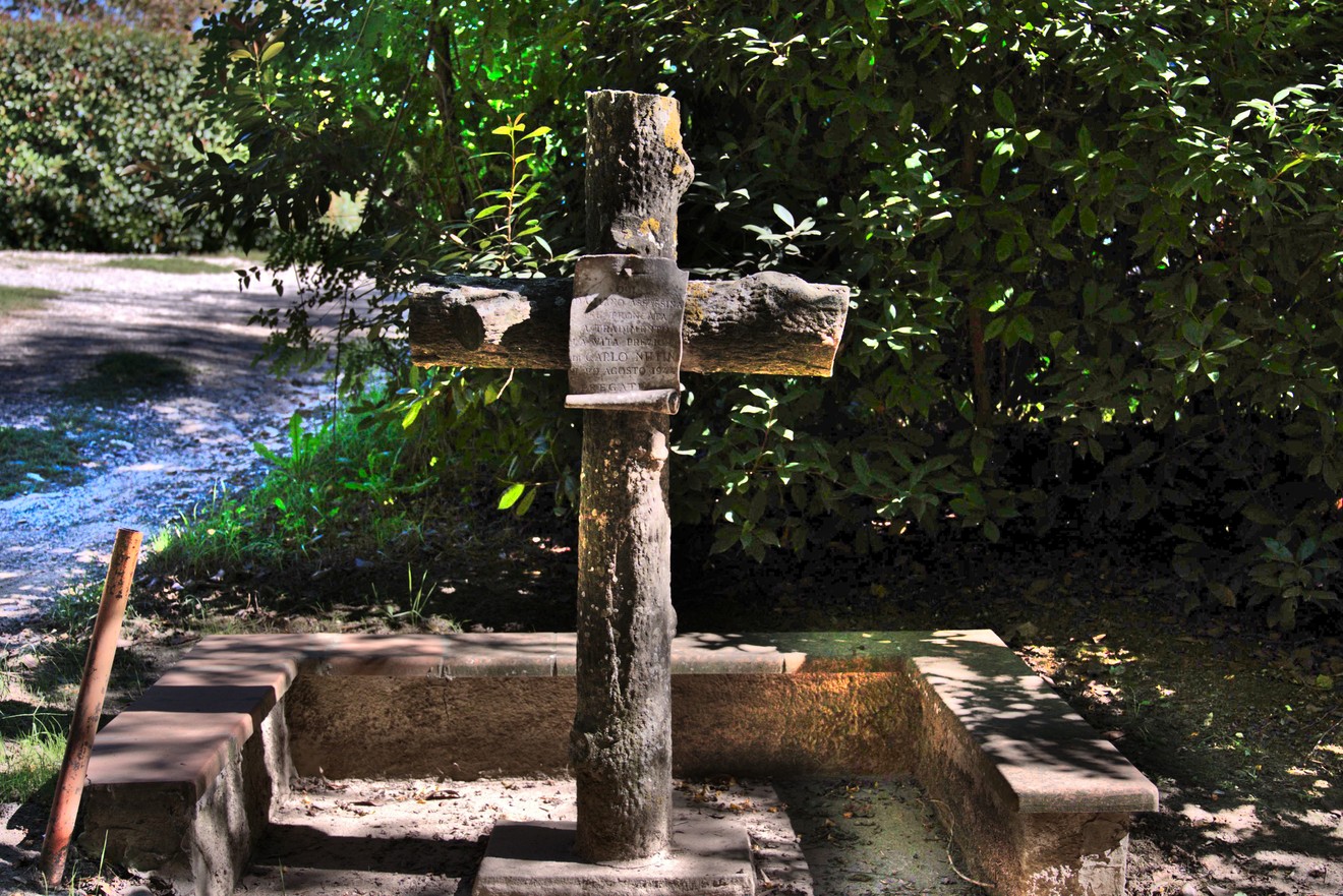 Croce in legno su base in pietra, con iscrizione, in una fontana immersa nel bosco.