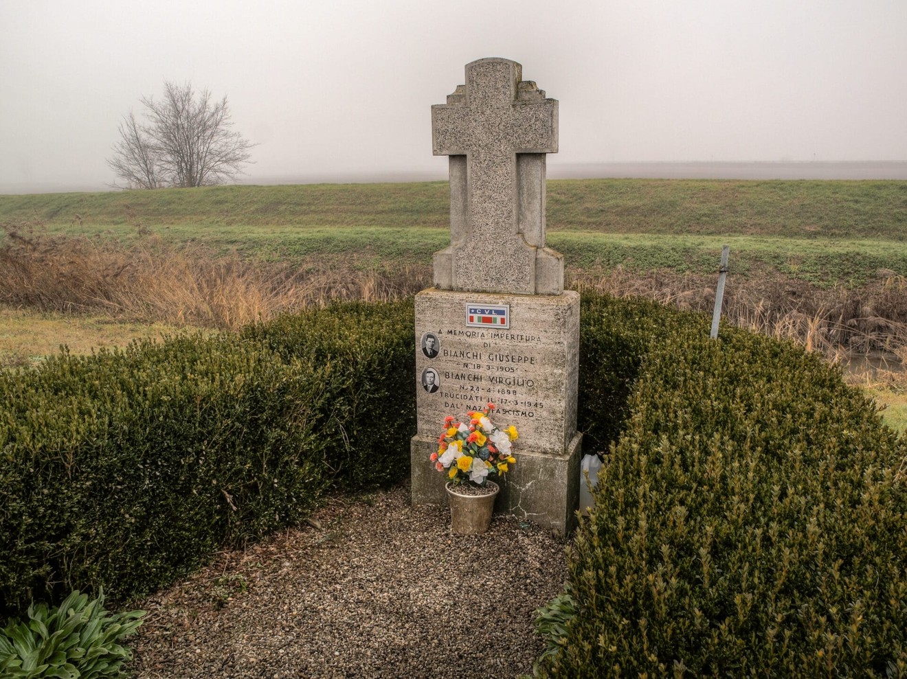 Cippo commemorativo con fiori, immerso in un paesaggio nebbioso.