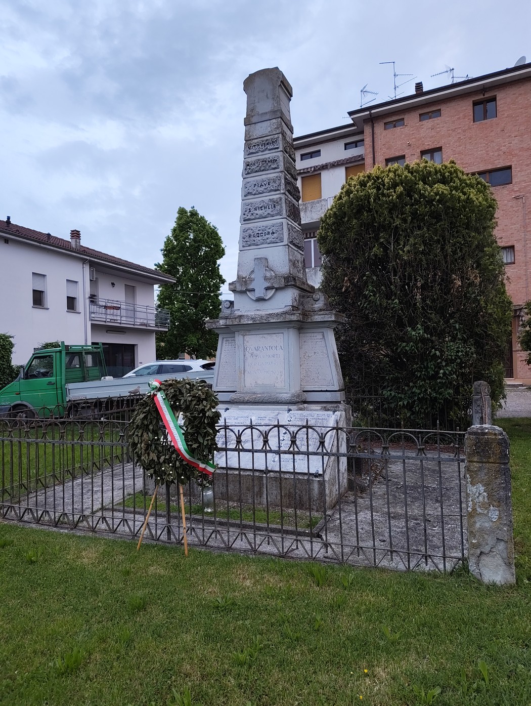 Monumento ai caduti di Quarantoli, circondato da verde e recinzione, in un contesto urbano.