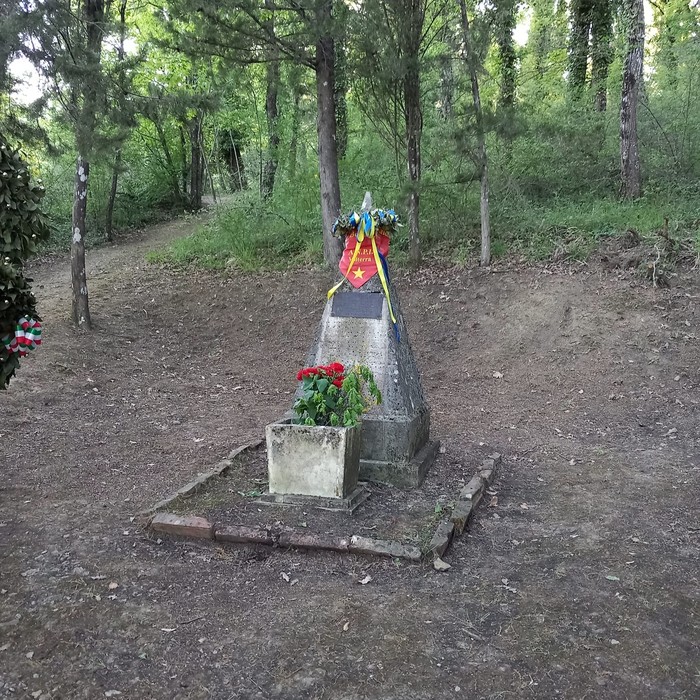 Cippo del podere La Casella circondato da vegetazione, con fiori e decorazioni commemorative.